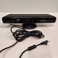 Front view of Xbox 360 Kinect sensor with cable coiled in front, showing glossy black finish and Xbox 360 branding.