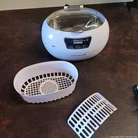 Full view of the white Fosmon ultrasonic jewelry cleaner with digital control panel, transparent hinged lid open, along with the included white plastic cleaning basket and divider on a brown surface.