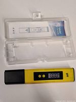 Yellow digital pH meter with black protective cap next to clear plastic case and instruction manual.