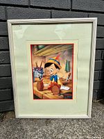 Full view of the framed lithograph showing Pinocchio and Jiminy Cricket in a workshop setting with a white frame and green mat.