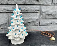 Front side of white ceramic Christmas tree with blue bulbs and attached power cord, placed against a gray stone wall.