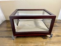 Rectangular display box with reddish-brown wooden frame, clear beveled glass on sides and top, supported by four metal elephant feet on wooden surface.