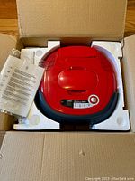 Top view of red Lentek Intelli-Vac robotic vacuum cleaner in original styrofoam packaging inside box, with instruction manual.