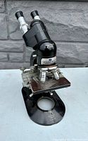 Front view of black metal compound microscope with binoculars and stage below.