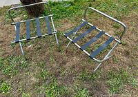 Pair of two vintage chrome luggage stands with black fabric straps displayed outdoors on grass.