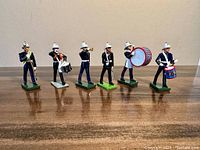 Full lineup of six vintage diecast Britains toy soldiers in military marching band attire standing on table, showing full figures and bases.
