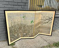 Four-panel folding screen leaning against a wall outdoors, showing full screen with hand-painted birds and blossoms on gold foil background in black lacquer frame.