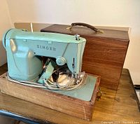 Singer 327K sewing machine mounted on wooden base with electrical cord, in turquoise color, next to original wooden carrying case on table.