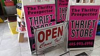 Two A-frame signs with 'The Thrifty Prospector THRIFT STORE NOW OPEN!' in pink and black colors, standing on floor.