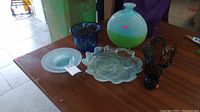 Five art glass items displayed on a wooden table: a large round vase primarily green and blue, a ruffled glass platter, two blue glass vases of different shapes, and a dark glass vase with artistic edges.