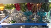 Collection of assorted colored hand blown glass stemware including green, red, amber goblets and blue cocktail glasses on shelf.