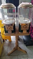 Front view of two gumball machines with clear containers, metal lids and coin slots mounted on wooden pedestal stand.