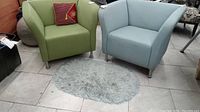 Two modern square club style arm chairs, one green and one blue, with a small oval gray shag rug in front of them
