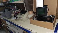 Shelf with various office supplies including Zebra thermal printer, clipboards, note pads, and a box with electronic devices