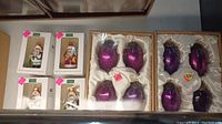 Four boxed hand decorated glass holiday ornaments next to two sets of four purple glass ornaments boxed in velvet/satin lined boxes.