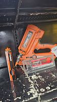 Orange Paslode cordless framing power nailer with nail cartridge and accessory case placed on a black surface.
