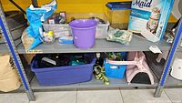 Shelf showing various pet supplies including containers, a purple bucket, plastic pet housings, cat food box and small pet cages.