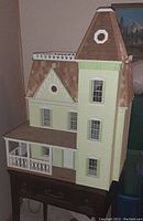 Full view of three-story wooden Victorian-style dollhouse painted light green with white trim and brown shingled roof, featuring front porch