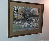 Frontal image of the framed printed portrait showing a woman walking among many white geese with trees and houses in the background, hung on a beige wall.