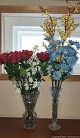 Two glass vases filled with artificial flowers, including red roses, white flowers, yellow and blue flowers, placed in front of a window.