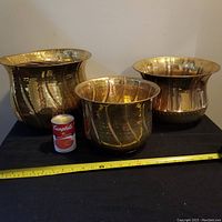 Three large brass planters of varying sizes and shapes displayed together with a Campbell's soup can for size reference, on a black surface against a plain wall.