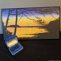 Wide photo showing argillite earrings in blue box in front of large decorative artwork reflecting natural landscape with sunset tones