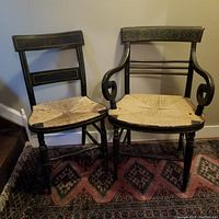 Two Hitchcock style antique dining chairs side by side against a wall on a patterned rug; one chair has scrolling arms, the other does not. Both have ebonized, hand painted frames and rush seats.
