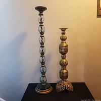 Two large vintage candle holders side by side against a neutral wall. The taller one has twisted glass or crystal segments with gold-tone base and top. The shorter one is brass with ornate openwork and raised design elements on the body and base.