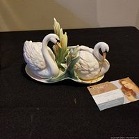 Pair of white porcelain swan salt and pepper shakers with green leafy accents, displayed on black cloth with attached tag from the Franz Collection.