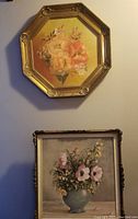 Two framed floral artworks hung on wall; one original oil painting with pink and white flowers in blue vase, the other a print of yellow and peach flowers in octagonal gold frame.