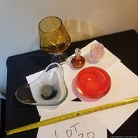 Overview of five glass items on black surface with tape measure and lot sign: clear smoke gray free form bowl, amber goblet, red bowl, pink vase, amber decanter