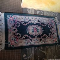 Full view of small wool rug showing dark green background with floral design in pink and blue, beige borders, and fringe ends.