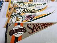 Four felt pennants featuring Oakland Athletics logos and San Francisco Giants graphics rolled and laid out with a mini Giants baseball bat.