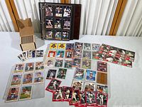Wide shot displaying binder with several pages of baseball cards, rows of loose cards spread on table, and a smaller cardboard box of baseball cards standing open to show many cards inside.