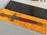 Full view of the decorative sword in its orange satin-lined display box, showing the blade sheathed and the detailed handle and scabbard fittings.