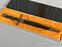 Full view of sword in scabbard in black box with yellow satin lining.