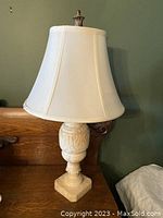 Full view of antique alabaster lamp with detailed carved base and white fabric shade against a green wall and wood furniture background.