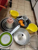 Photo showing an overview of mixing bowls, yellow pitchers, a Bundt pan, Tupperware shaker, collapsible strainer, measuring spoons, and mesh strainer on tiled floor.