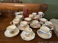 Wide view of assorted commemorative china cups and mugs arranged on wooden table, inclusive of saucers and detailed designs