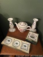Overview showing two white ceramic candlesticks with floral decorations, a matching ceramic teapot with lid, a boxed coaster set, and a boxed small decorative frame all decorated with Portmeirion Botanic Garden floral motifs.