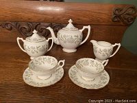 Full tea set arranged on wooden surface consisting of teapot, lidded sugar bowl, creamer, two cups and two saucers, showing floral pattern and gold trim