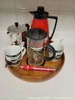 Lot of coffee accessories including Bodum French press, espresso pot, red thermos, two ceramic mugs, and espresso tamper displayed on a wooden tray
