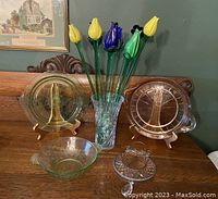 Glass flowers arranged in clear textured vase with various glassware pieces including round etched plates, green glass bowl, and glass trivet on wooden surface under framed picture.