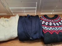 Three folded men's sweaters: cream cable knit pullover, navy cardigan, black/red patterned pullover
