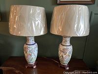 Pair of porcelain lamps with plastic-covered beige fabric shades, brass tops, and wooden bases placed on a wooden surface against a green wall.