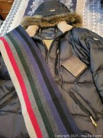 Black Point Zero winter coat laid flat with multicolor striped knit scarf and brown leather wallet on top.