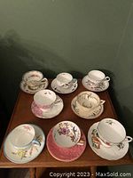 Top view showing 8 different china teacups and matching saucers arranged on a wooden table, each with unique delicate floral or decorative patterns, some with gold trim.