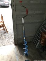 Full view of the 6-inch manual ice auger with red handle against a wooden floor and metal wall.