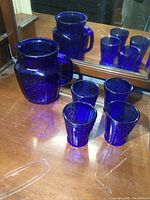 Photo showing cobalt blue glass pitcher and four matching glasses reflected in mirror, placed on wooden surface.