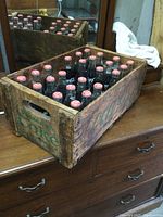 Front and top view of wooden Coca Cola crate filled with 24 full glass bottles with red caps, showing the crate's aged wood and printed logo.